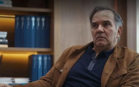 Em cena de Travessia, Humberto Martins está com a expressão de preocupação:ele usa blusa azul marinho com um casaco caramelo por cima. Ao fundo, uma estante com livros