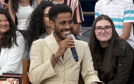 De blazer bege, Ícaro Silva sorri com o microfone nas mãos no palco do Altas Horas