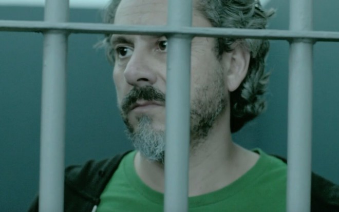 O ator Alexandre Nero com expressão séria, atrás das grades em cena de Império