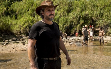 José Alfredo (Alexandre Nero) está com chapéu na cabeça e com os pés na beira de um rio no garimpo em cena de Império