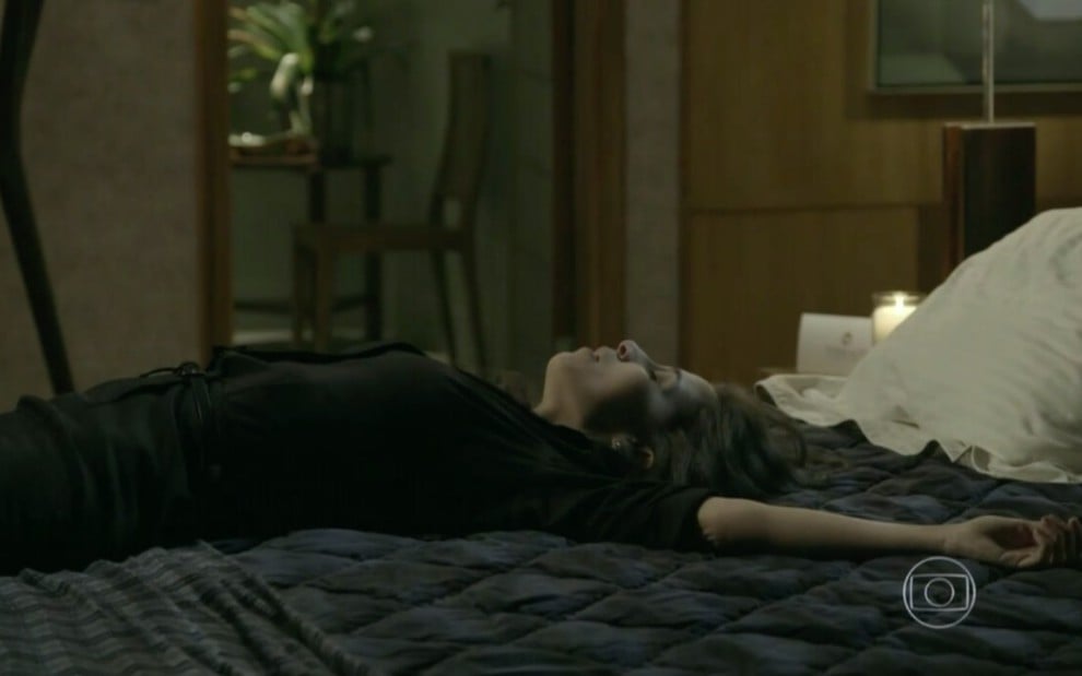 A atriz Marjorie Estiano toda de preto, deitada numa cama de braços abertos e olhos fechados, em cena de Império