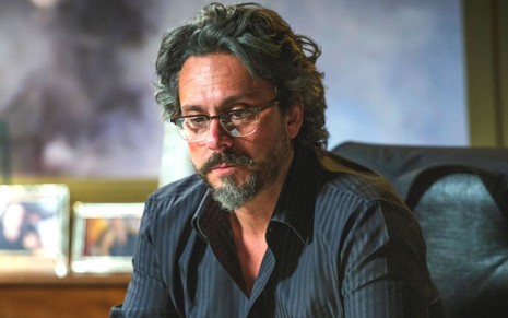 O personagem José Alfredo (Alexandra Nero) está de óculos e sentado em uma cadeira em cena de Império, novela da Globo