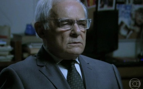 O ator Othon Bastos com expressão séria, óculos, terno e gravata em cena de Império