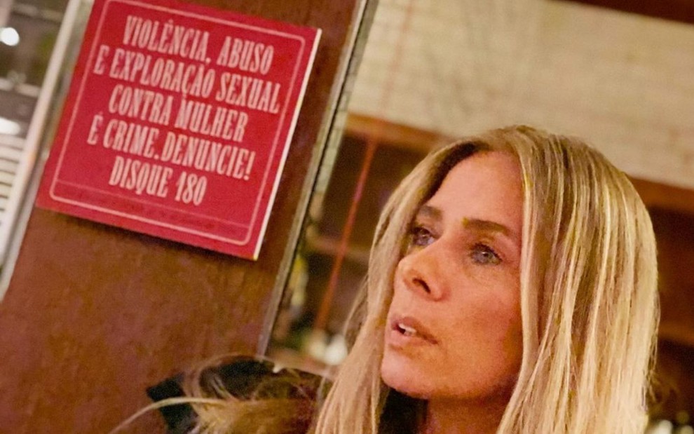 A apresentadora Adriane Galisteu em foto publicada no Instagram em que aparece com uma placa ao fundo incentivando denúncias de violência contra a mulher