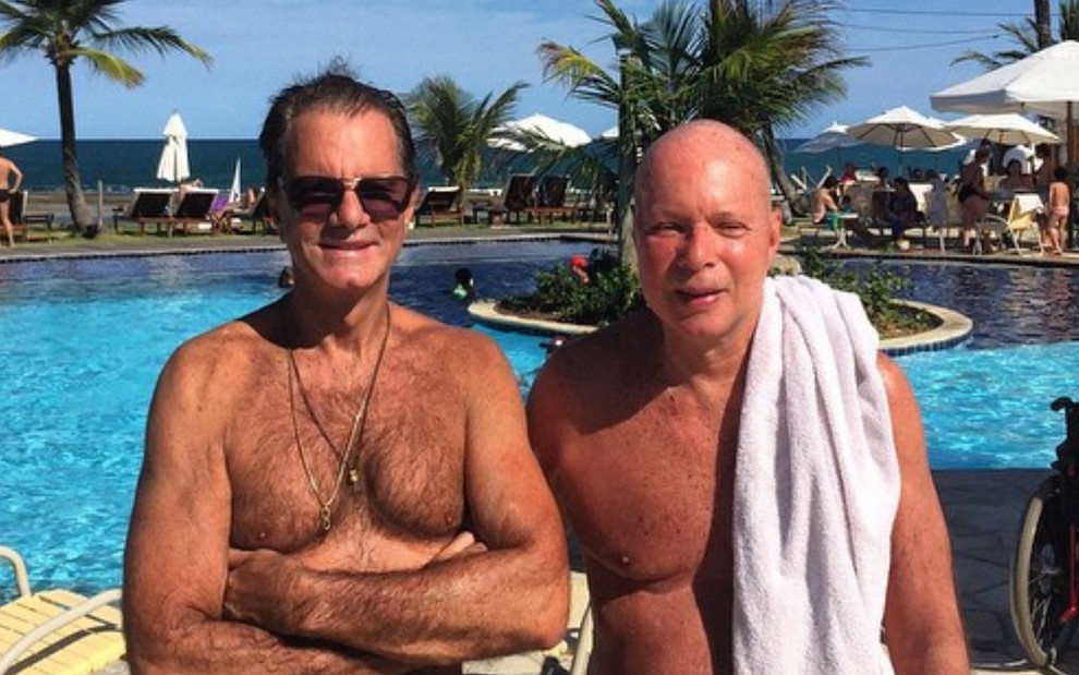 Foto do casal Edgar Moura Brasil e Gilberto Braga em que os dois aparecem sem camisa à beira de uma piscina, que está ao fundo