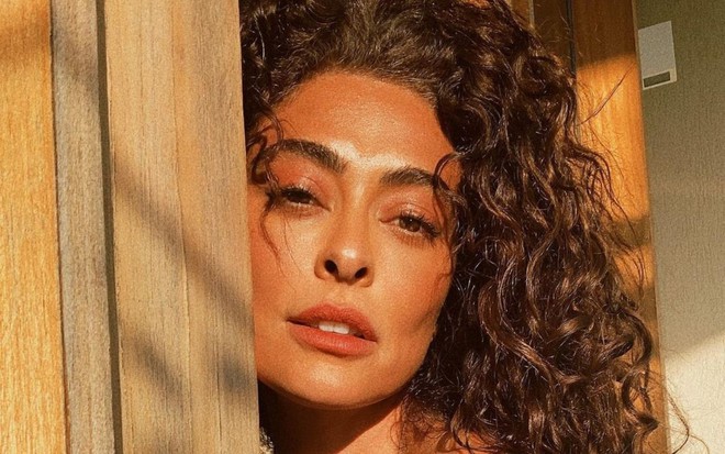 Juliana Paes está de cabelo solto e cacheado; ela está encostada em uma porta