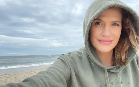 Com um moletom e capuz, Kristy Swanson tira selfie em uma praia