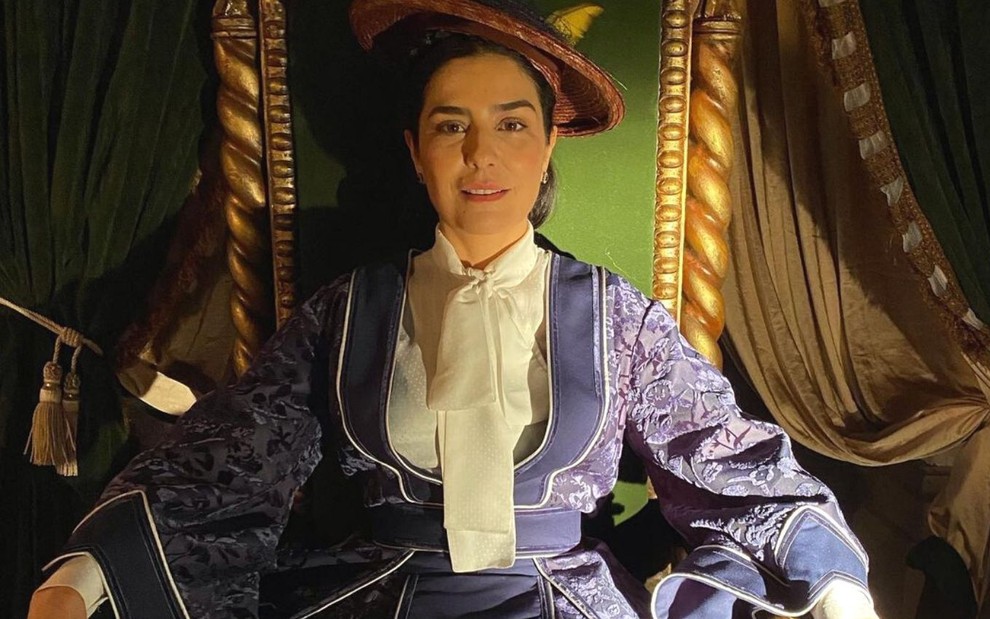 Leticia Sabatella está caracterizada como Teresa Cristina, ela usa vestido de época e chapéu. A atriz está sentada em um trono nos bastidores da novela Nos Tempos do Imperador