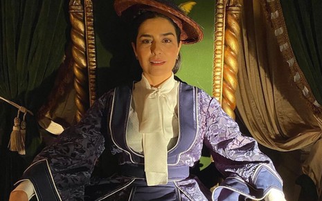 Leticia Sabatella está caracterizada como Teresa Cristina, ela usa vestido de época e chapéu. A atriz está sentada em um trono nos bastidores da novela Nos Tempos do Imperador