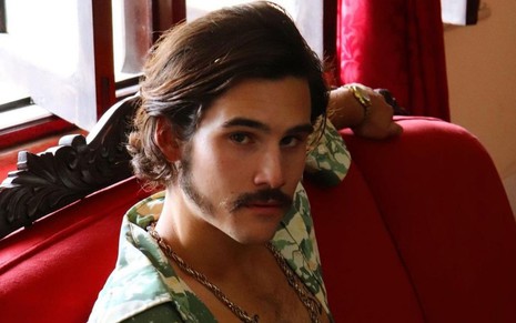 Nicolas Prattes caracterizado como o mafioso Giovanni Nicola em série da Globo: ator está com blusa aberta no peito, usa colar e está com o cabelo no ombro e bigode