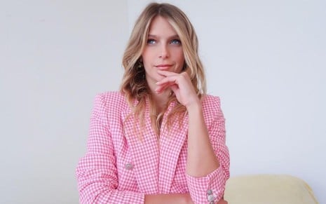 Isabella Santoni veste uma camisa rosa e faz uma pose sentada com a mão esquerda apoiada no queixo e a mão direita abraçando a barriga
