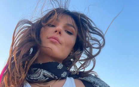 Isis Valverde faz selfie de baixo para cima; ela está com os cabelos ao vento, veste regata branca e usa bandana preta no pescoço