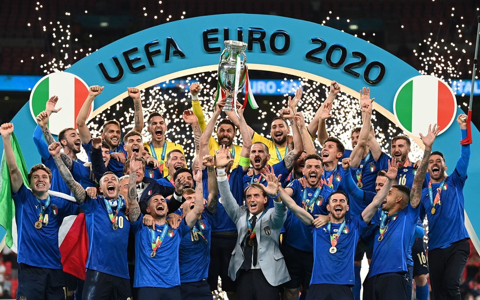Jogadores da Itália levantam o troféu da Eurocopa, em um pódio azul. Todos eles usam a camisa azul da seleção italiana, com exceção dos goleiros, que estão de amarelo