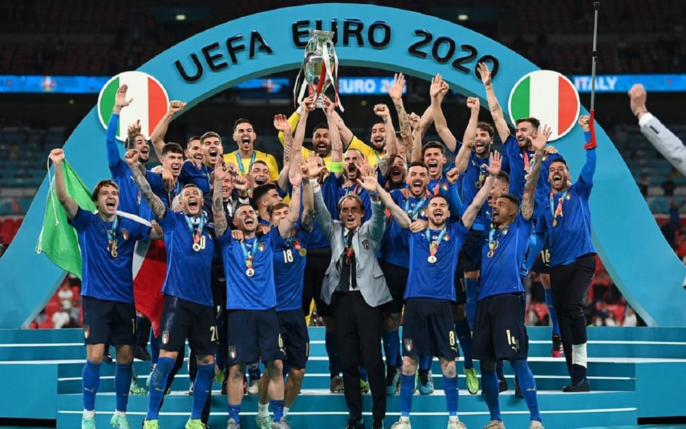 Jogadores da Itália levantam o troféu da Eurocopa, em um pódio azul. Todos eles usam a camisa azul da seleção italiana, com exceção dos goleiros, que estão de amarelo