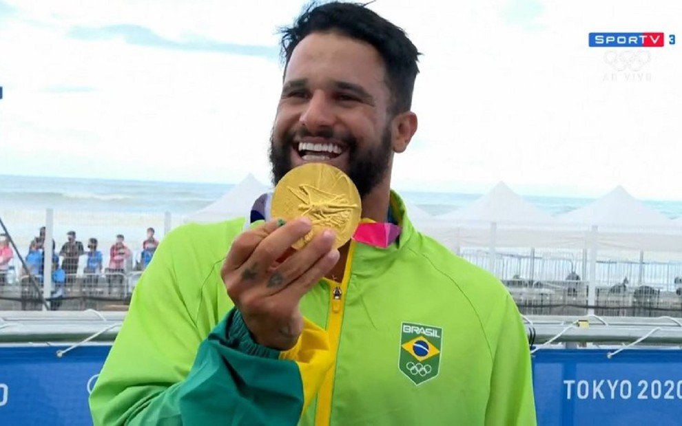 Ítalo Ferreira com um casaco verde e amarelo do time Brasil nos Jogos Olímpicos, mostrando e sorrindo a medalha de ouro que ganhou em Tóquio