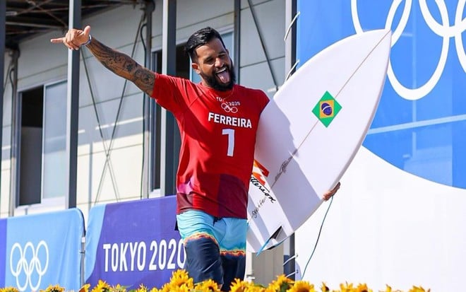 Italo Ferreira com uma camiseta vermelha e bermuda, segurando uma prancha, acenando com uma mão e mostrando a língua