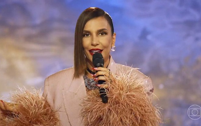 Ivete Sangalo com um vestido bege e sorrindo para a câmera no cenário do The Masked Singer Brasil. O fundo é azul.