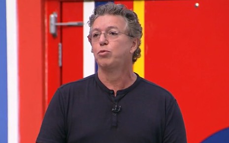 Foto de Boninho; ele veste camiseta preta e está na frente de uma parede vermelha