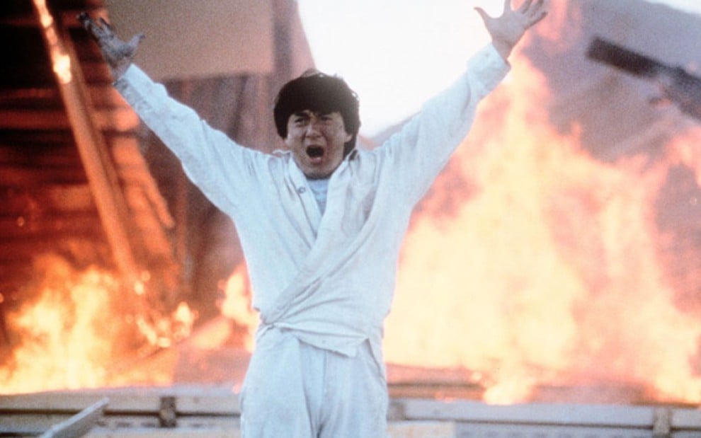 Jackie Chan vestindo roupas brancas, com os braços para cima e a boca aberta; atrás, o ambiente pega fogo