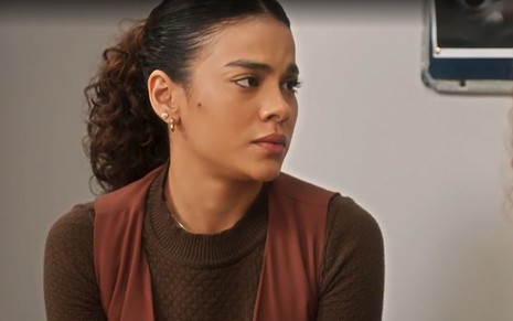 Em cena de Vai na Fé, Bella Campos está conversando com alguém com a expressão de tristeza; ela veste roupa marrom e está de rabo de cavalo
