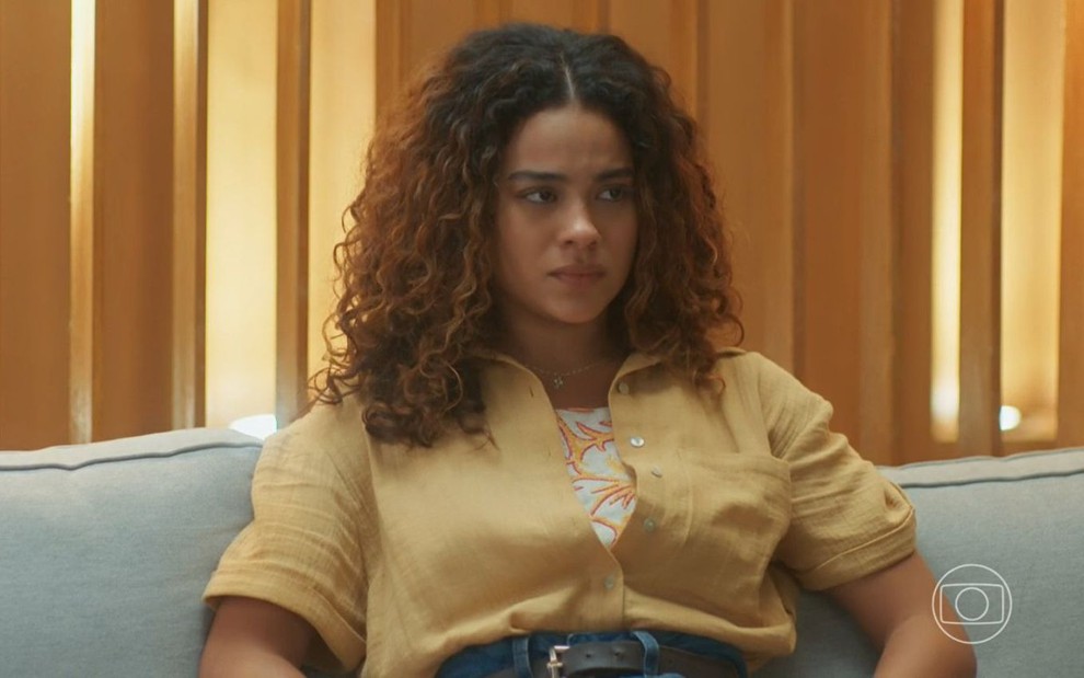 Em cena de Vai na Fé, Bella Campos está sentada em um sofá, desolada: ela usa jeans, camisa de botão amarela e cabelos soltos