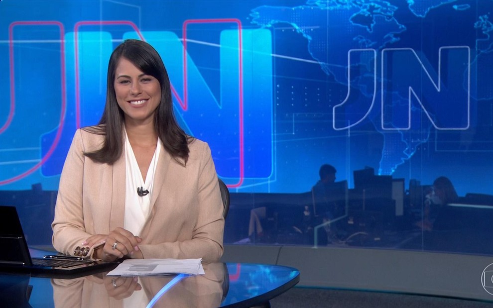 Jéssica Senra, âncora da Globo, no cenário totalmente azul do Jornal Nacional. Ela sorri e usa um blazer rosa com uma camisa branca por dentro.