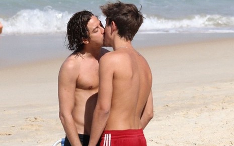 O ator Jesuita Barbosa e o designer gráfico Cícero Ibeiro; eles estão na praia, de frente ao mar, se beijando com as mãos dadas