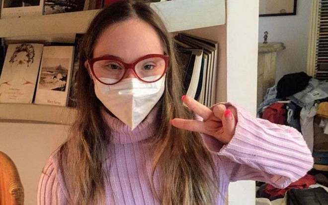 A atriz Joana Mocarzel faz sinal de V com as mãos, usa máscara e óculos de grau, além de blusa de lã lilás
