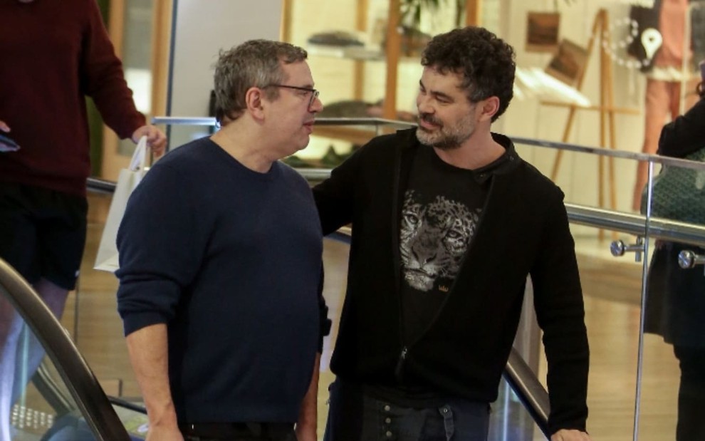 João Emanuel Carneiro e Carmo Dalla Vecchia encaram um ao outro; ator e novelista dão um leve sorriso em meio a escada rolante do Village Mall