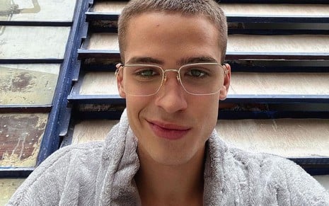 João Guilherme usa óculos, tem o cabelo raspado e dá um sorriso de lado