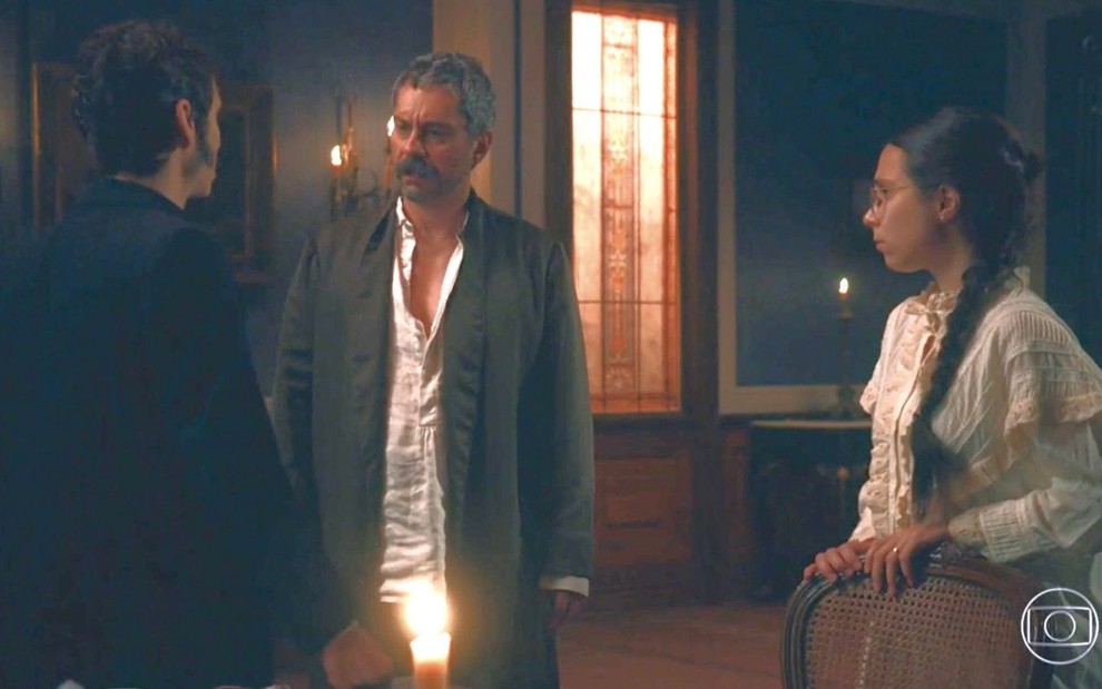 De costas, Nélio (João Pedro Zappa) olha para Tonico (Alexandre Nero); Dolores (Daphne Bozaski) observa os dois em cena de Nos Tempos do Imperador
