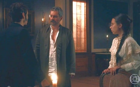 De costas, Nélio (João Pedro Zappa) olha para Tonico (Alexandre Nero); Dolores (Daphne Bozaski) observa os dois em cena de Nos Tempos do Imperador