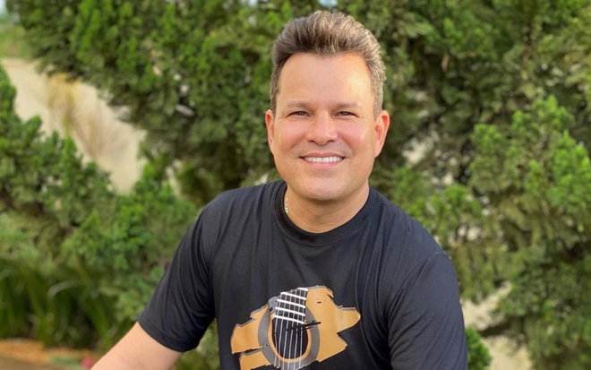 João Neto está sentado e sorri para foto; cantor usa calça e camiseta preta
