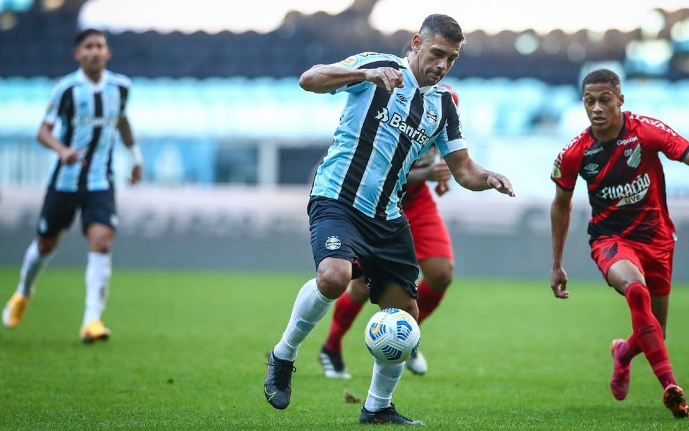 Diego Souza com camisa azul, preta e branca, calção preto e meião branco do Grêmio domina a bola com pé direito levantado sendo observado por um marcador de uniforme vermelho