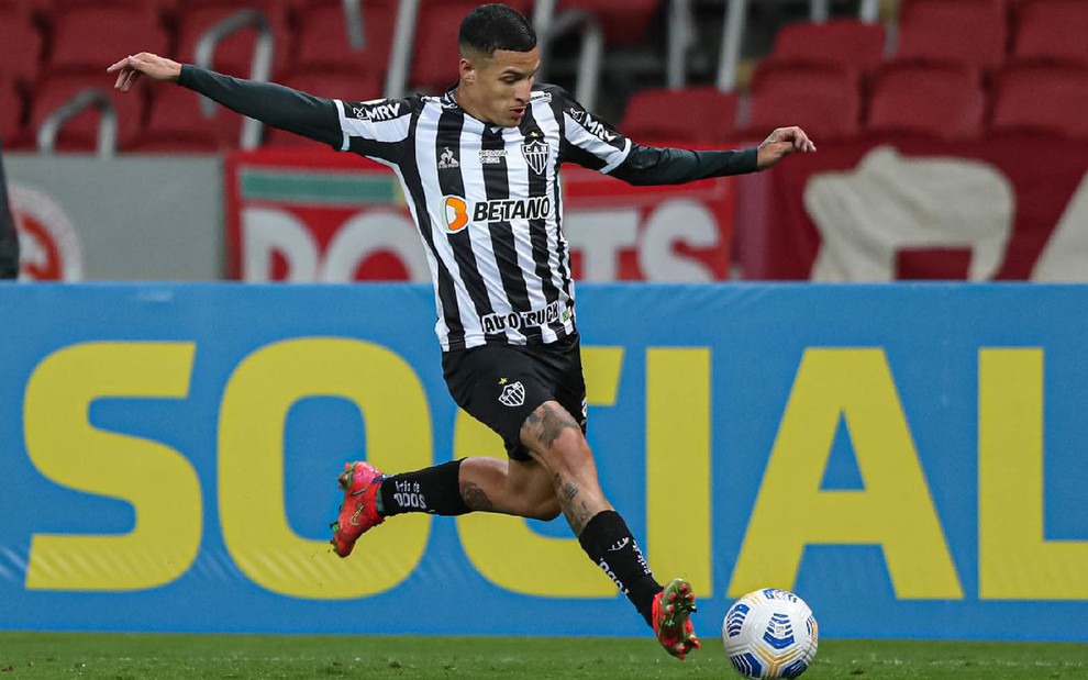 Guilherme Arana com meião e calção preto e camisa preta e branca do Atlético-MG em movimento para chutar a bola com o pé esquerdo em partida