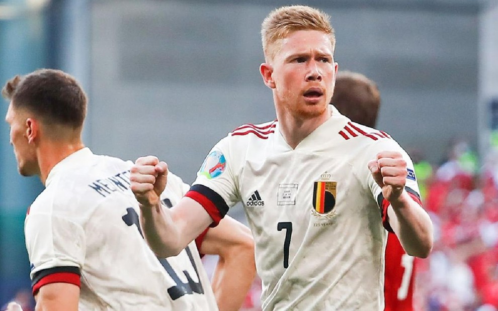 Kevin De Bruyne com uniforme branco da Bélgica celebra gol com punhos cerrados e olhando para o lado direito em campo