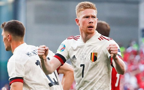 Kevin De Bruyne com uniforme branco da Bélgica celebra gol com punhos cerrados e olhando para o lado direito em campo