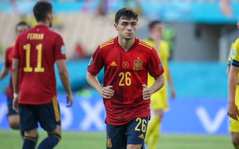 Pedri com calção azul e camisa vermelha da seleção da Espanha corre com os braços juntos na cintura com outro jogador da Espanha no lado esquerdo desfocado no fundo