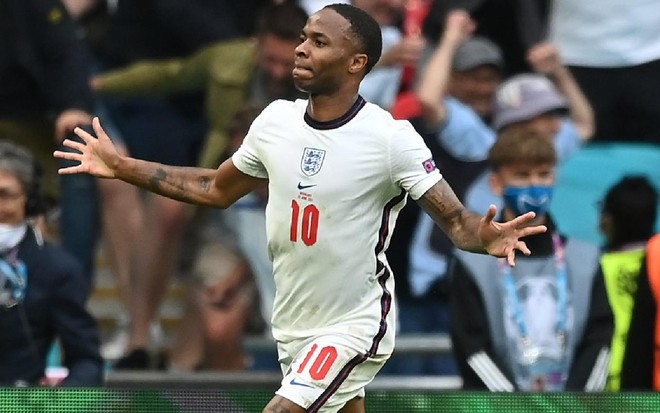 Raheem Sterling com uniforme todo branco da Inglaterra com as mãos abertas na altura da cintura correndo para o lado esquerdo em celebração de gol em partida