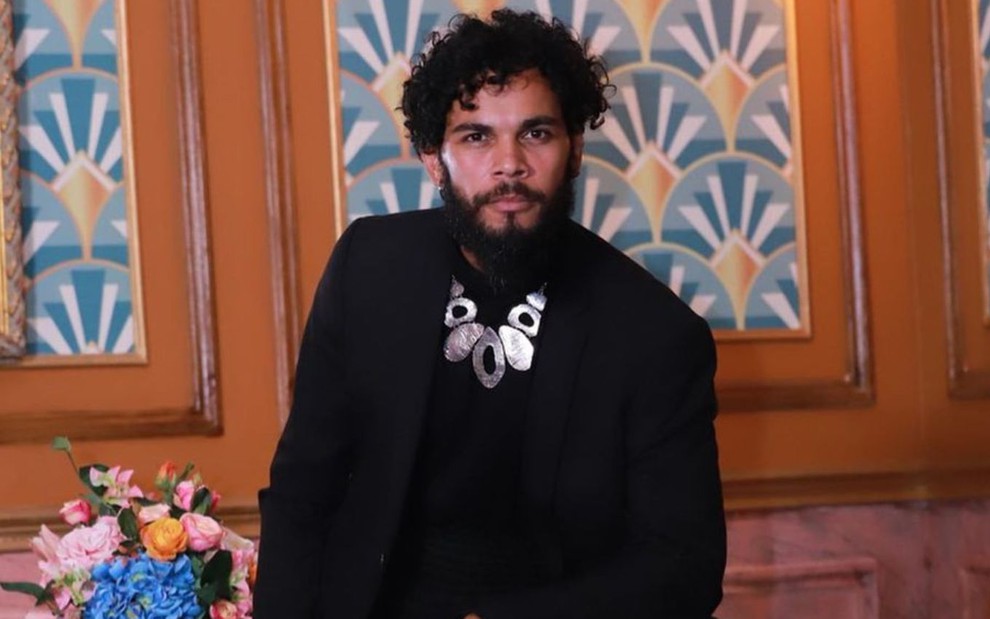 O ator Jorge Florêncio posa para foto de terno, camisa preta, colar prateado, expressão séria