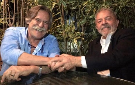O ator José de Abreu e o presidente eleito Lula, em foto de 2017; eles estão sentados a uma mesa, dando as mãos um ao outro e sorrindo para a foto