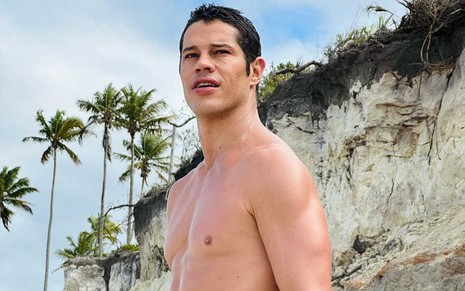 O ator José Loreto sem camisa, com expressão séria, em foto tirada de baixo para cima em cenário de praia