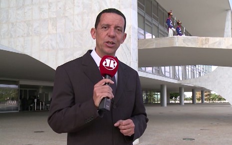 José Maria Trindade com um terno marrom, em frente ao Palácio do Planalto, com o microfone da Jovem Pan nas mãos