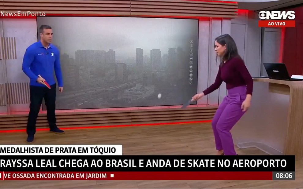 Alexandre Oliveira usa uma blusa azul e camisa preta, enquanto Júlia Duailibi usa uma blusa vinho e calça roxa. Ambos jogam tênis de mesa no estúdio da GloboNews