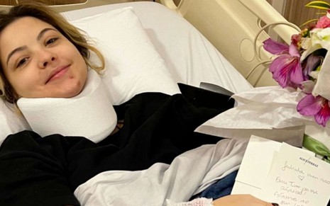 A atriz Júlia Gomes deitada em um leito de hospital com o pescoço imobilizado