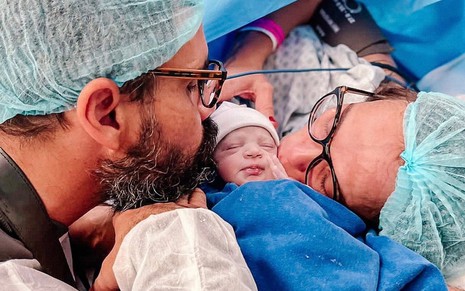 Juliano Cazarré com a filha Maria Guilhermina no colo diante de uma janela no hospital