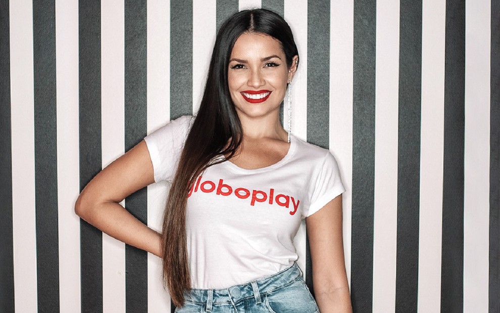 Juliette Freire com uma camisa branca que conta com a logomarca do Globoplay. O fundo é listrado em preto e branco e ela sorri para a câmera