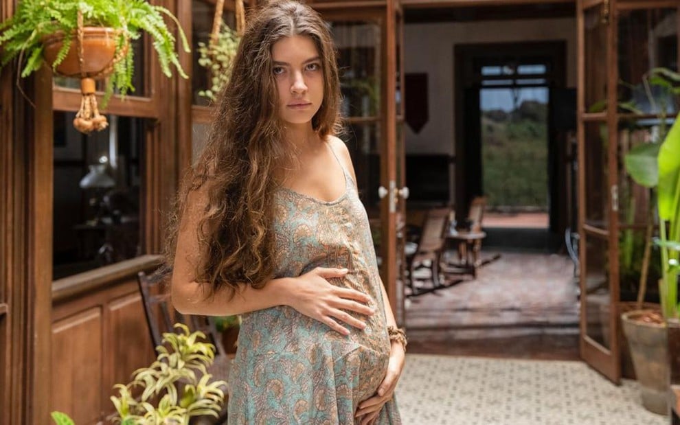 Caracterizada como sua personagem em Pantanal, Alanis Guillen está segurando a barriga de grávida