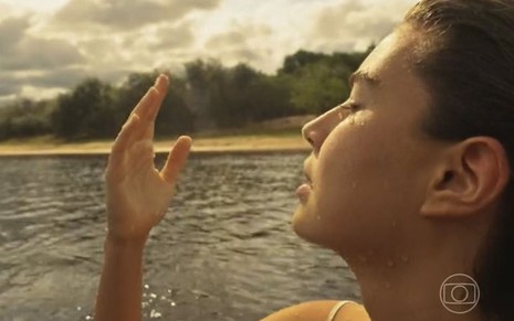 A atriz Alanis Guillen como Juma em Pantanal; ela está dentro do rio, sendo batizada, de olhos fechados, com a mão levantada a sua frente