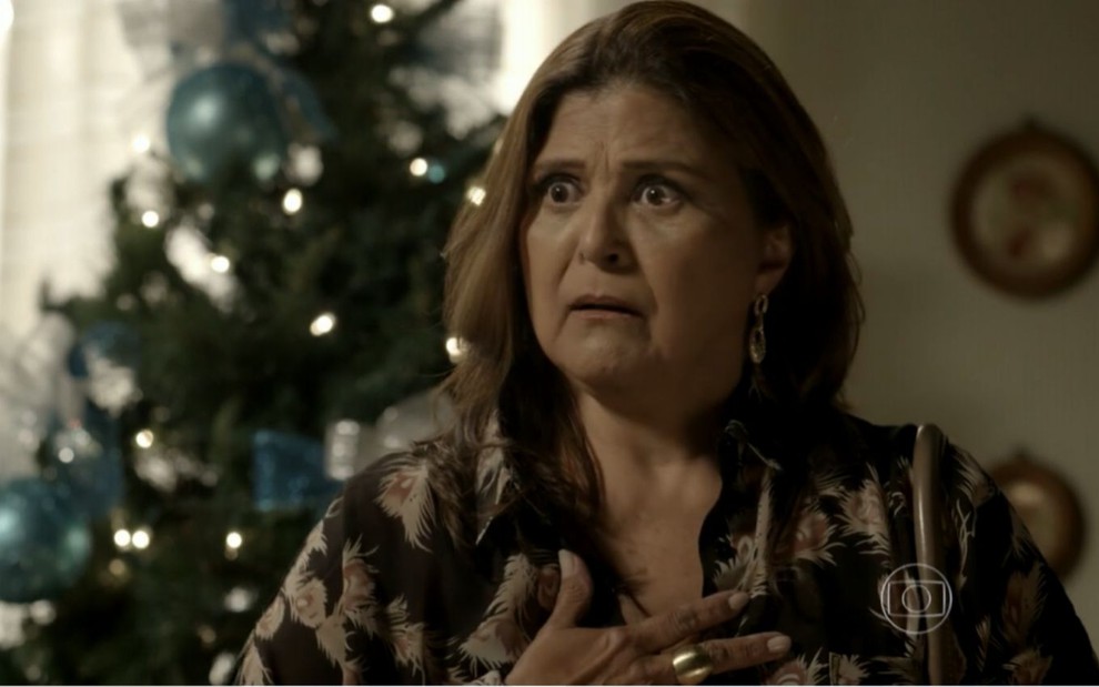 Jurema (Elizangela) está com a mão no peito e demonstra surpresa na casa de Cora (Marjorie Estiano); a sala está decorada com peças de Natal